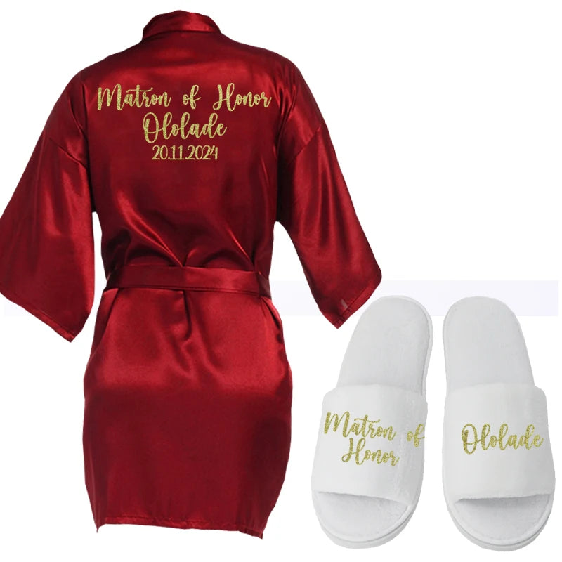Red satin robe with "Matron of Honor Ololade 20.11.2024" in gold script on back, paired with white slippers embroidered with "Matron of Honor" and "Ololade" in matching gold text.