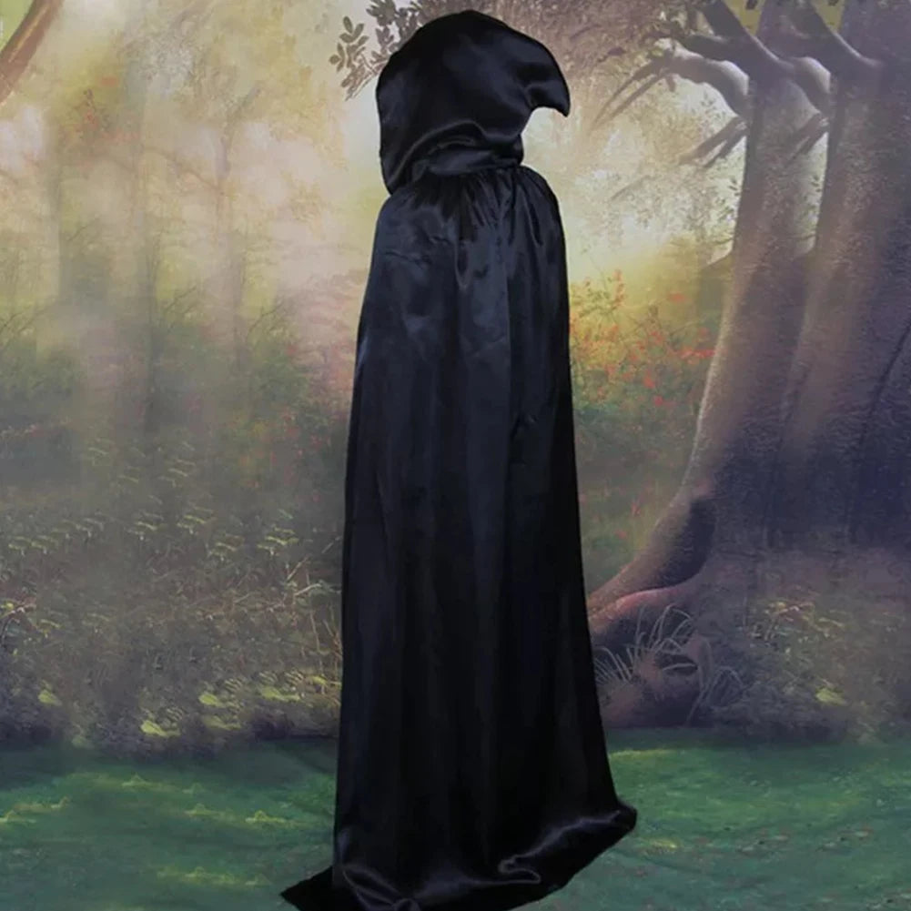 A person in a long black hooded cloak stands in a misty forest, surrounded by tall trees and soft green foliage. The atmosphere is mysterious and slightly ominous.