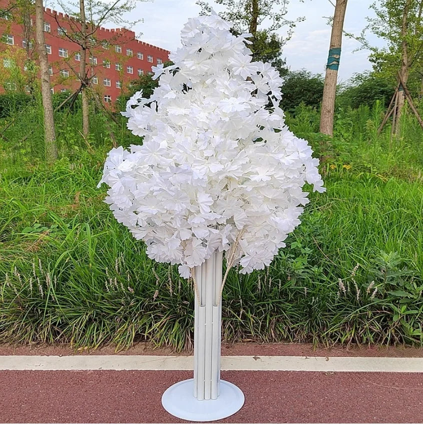 A decorative tree-like arrangement with white artificial blossoms on a white cylindrical stand, set on a path with greenery and a red brick building in the background.
