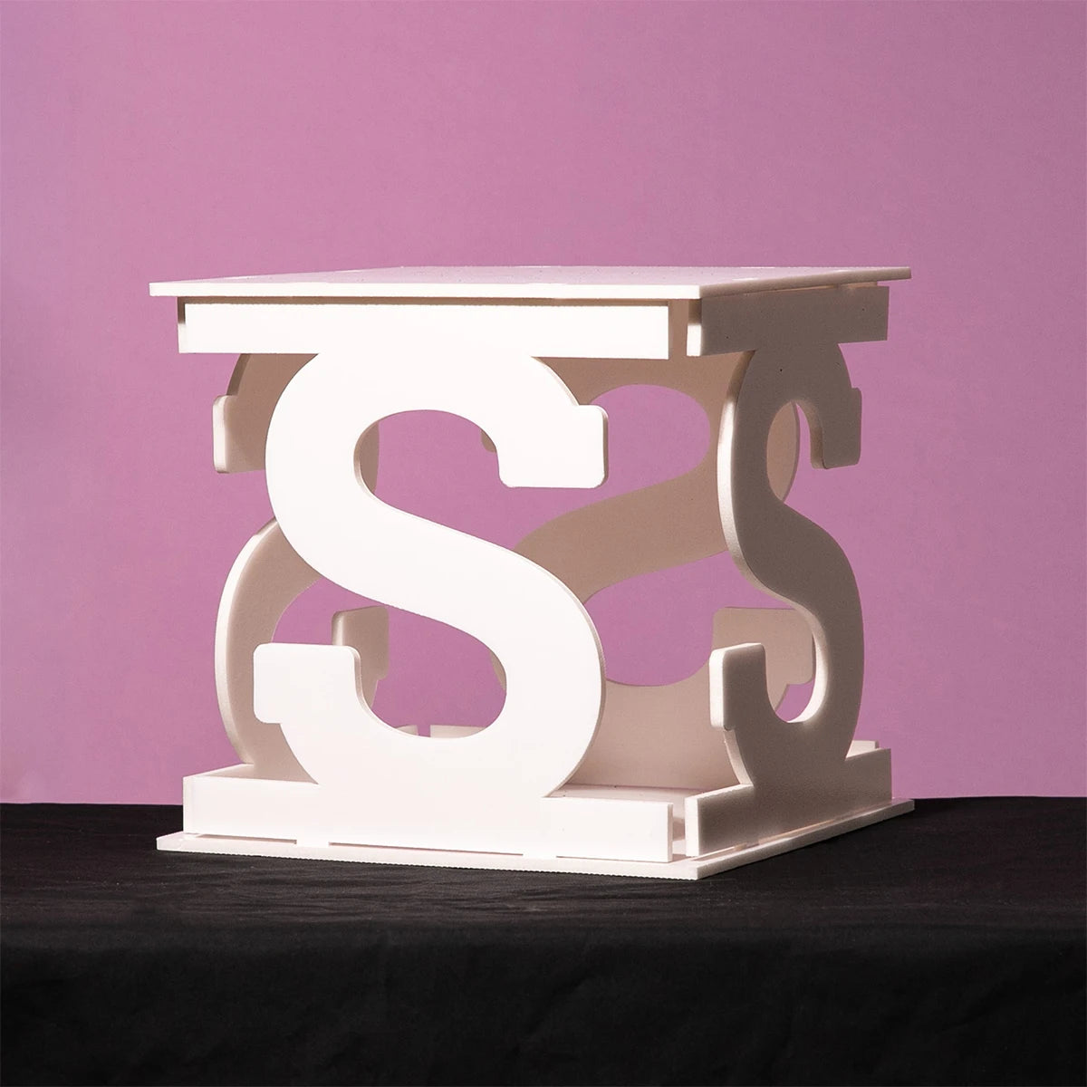 White stool made of interlocking "S" shapes, placed on a dark surface against a pink background. The design appears modern and artistic.