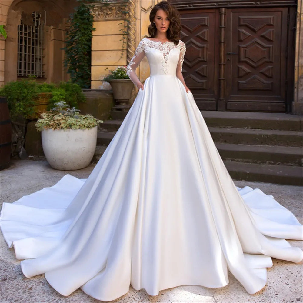 A woman in a white wedding dress with lace sleeves stands outdoors. The dress has a full skirt with a long train. There are stone steps and potted plants in the background.