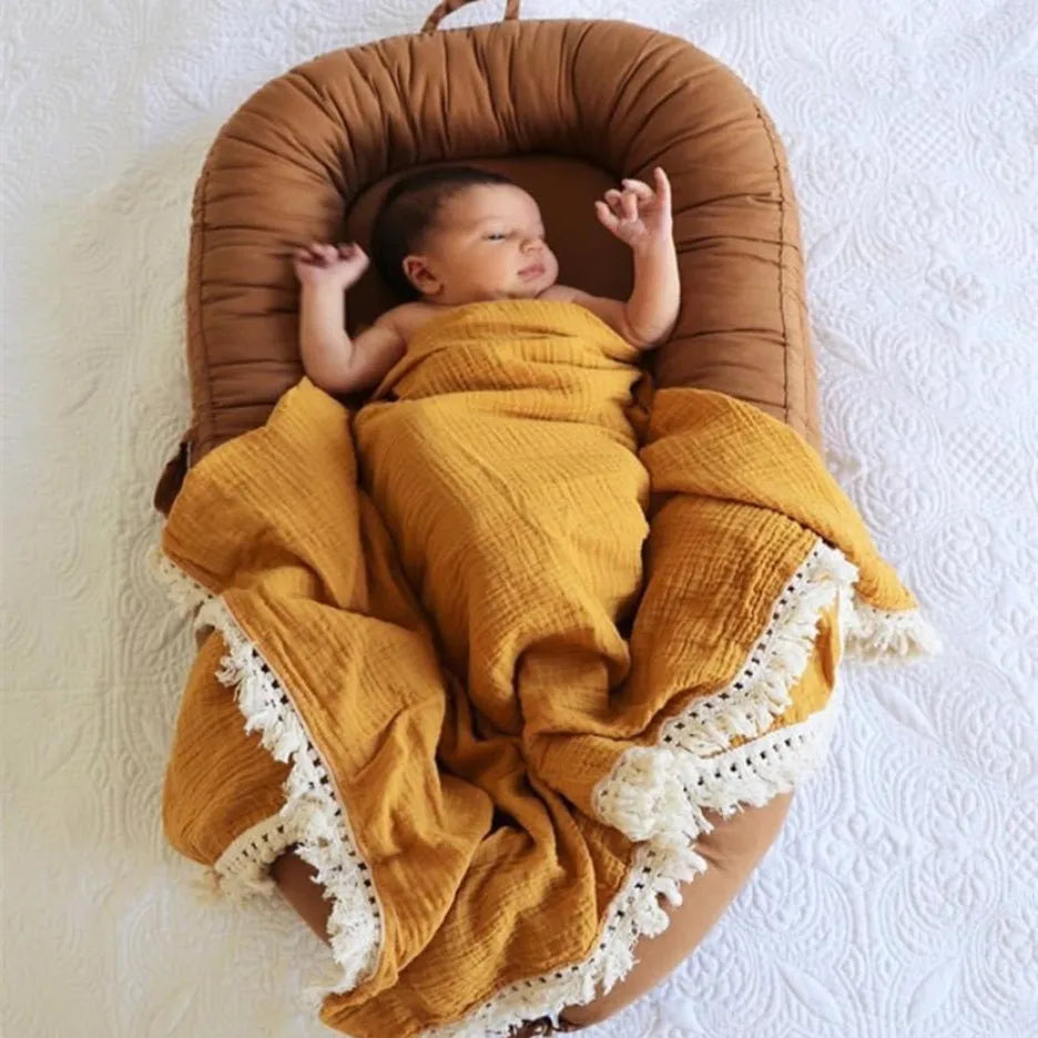 A baby sleeps in a cozy, brown nest with arms raised, wrapped in a mustard-colored blanket with white lace edges, on a textured white surface.