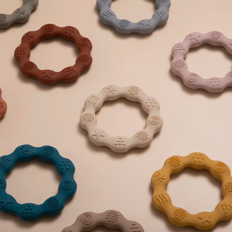 Colorful, textured silicone teething rings with wavy edges arranged in a scattered pattern on a neutral background. Rings feature various embossed patterns for sensory stimulation.
