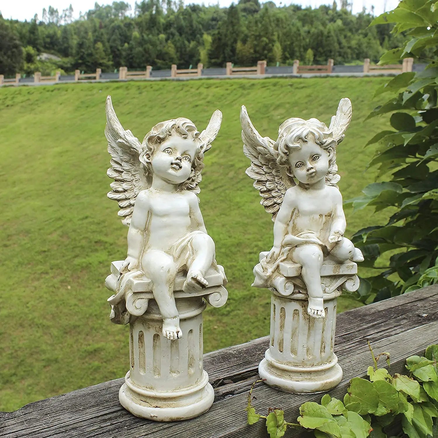 Two stone cherub statues with wings sit on ornate pedestals. They are positioned on a wooden ledge with lush green lawns and trees in the background.