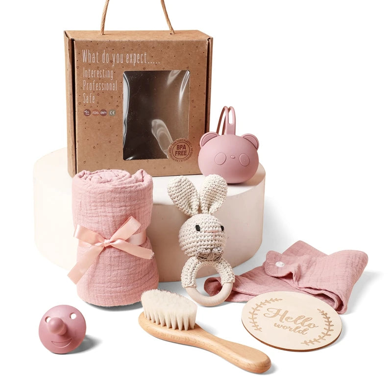 A baby gift set including a pink swaddle, bunny rattle, brush, pink silicone pacifier, bear-shaped teether, pink cloth, and a "Hello World" wooden sign, all with eco-friendly packaging.