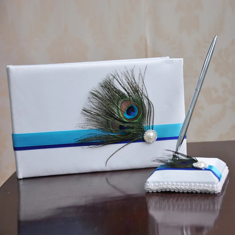 White guest book with peacock feather and a ribbon, paired with a pen on a cushioned base, adorned with matching colors and pearl details, placed on a wooden surface.