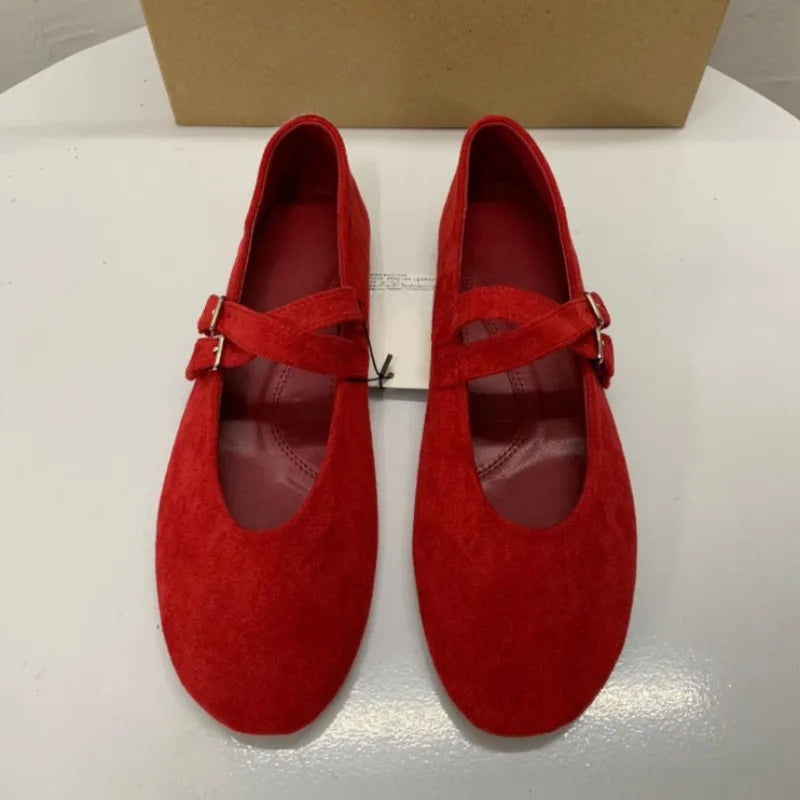 Pair of red suede ballet flats with cross straps and silver buckles, displayed on a white surface with a brown box in the background.