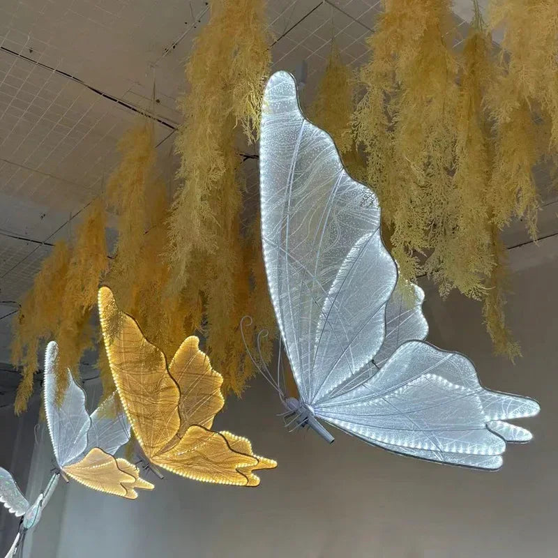Glowing butterfly sculptures, in white and yellow, hang amid wispy dried foliage from a ceiling grid, creating an ethereal atmosphere in the room.
