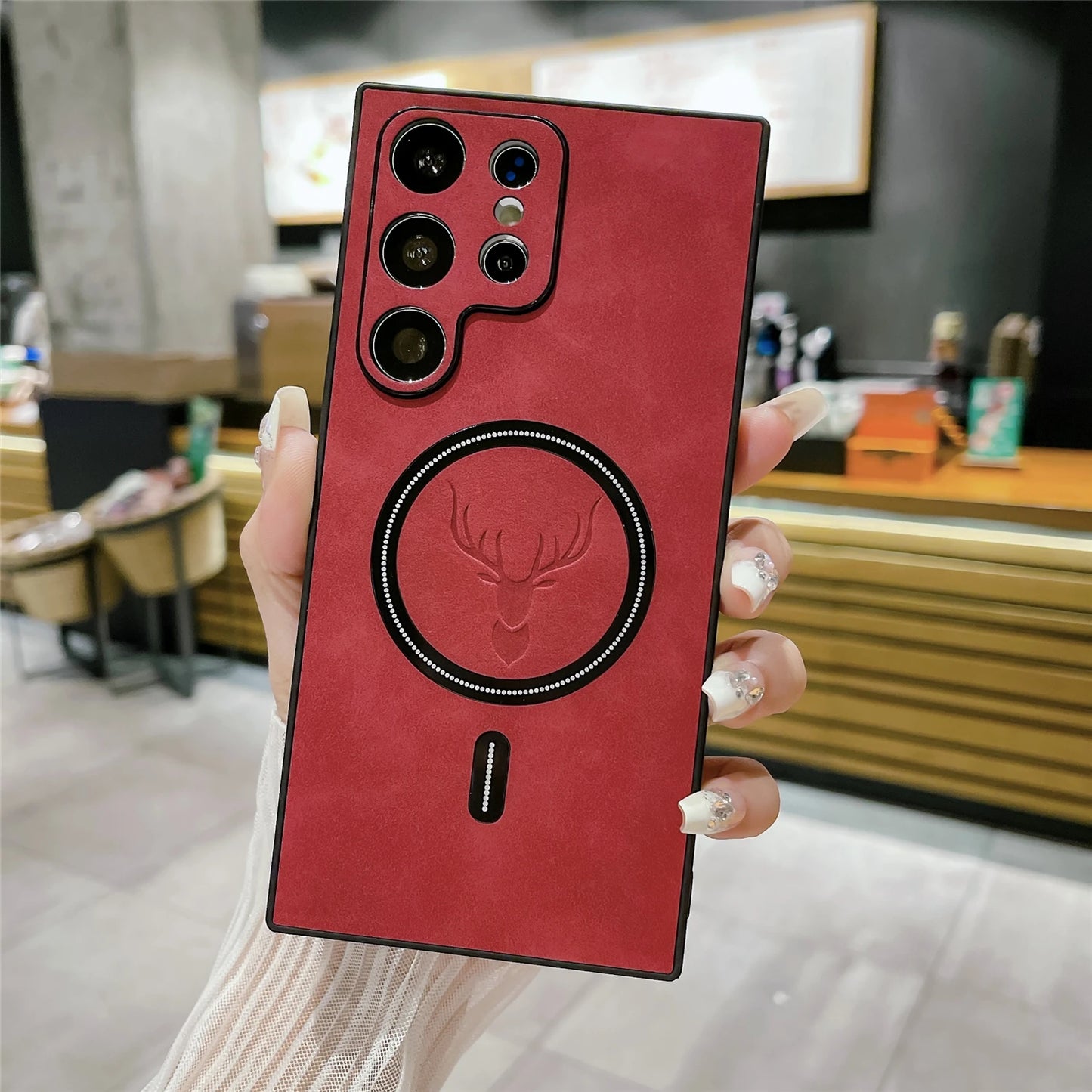 A hand holds a smartphone with a red leather case featuring a raised deer head design and multiple camera lenses. The background shows a blurred indoor setting.