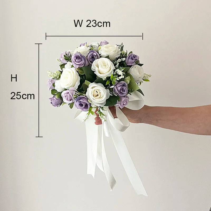 A person holds a bouquet of white and lavender roses measuring 23cm wide and 25cm high. The bouquet features white ribbons and small green leaves against a neutral background.