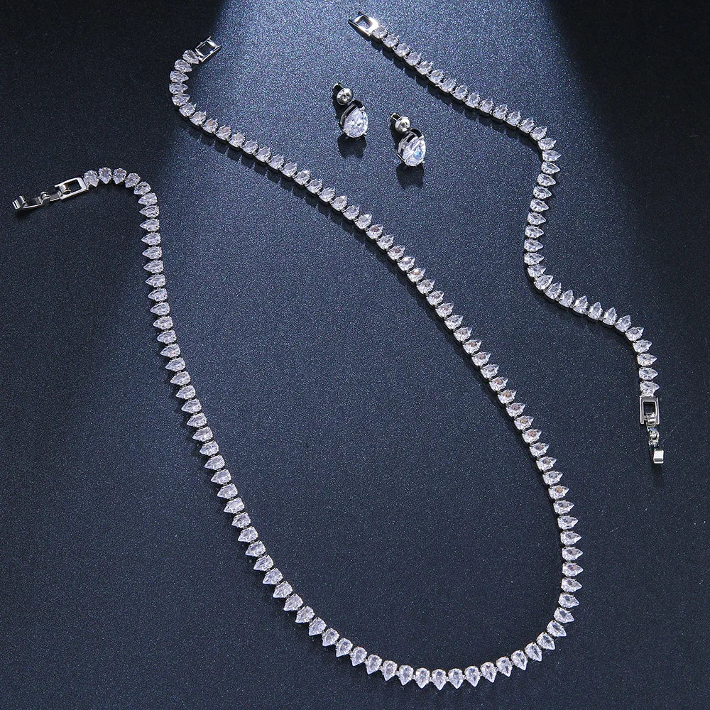 Jewelry set featuring a sparkling necklace and bracelet, both adorned with pear-shaped diamond-like stones. Accompanied by matching oval stud earrings on a dark background.