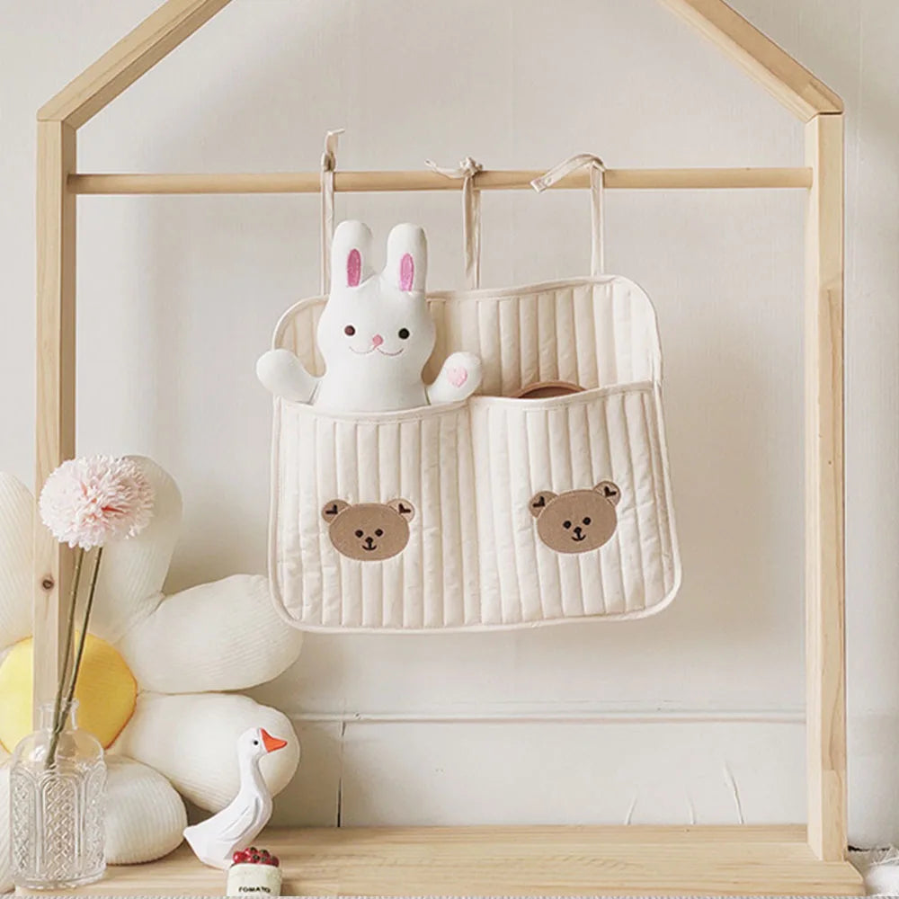 Hanging fabric organizer with bear faces, holding a white rabbit plush. A large flower cushion, glass vase with a pink flower, and a small goose figurine are on a wooden shelf.