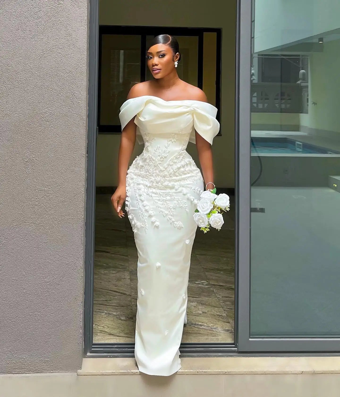 Woman in elegant off-shoulder white gown with floral details, holding white roses, stands in a doorway. Hair is sleek and styled, and she wears matching earrings.