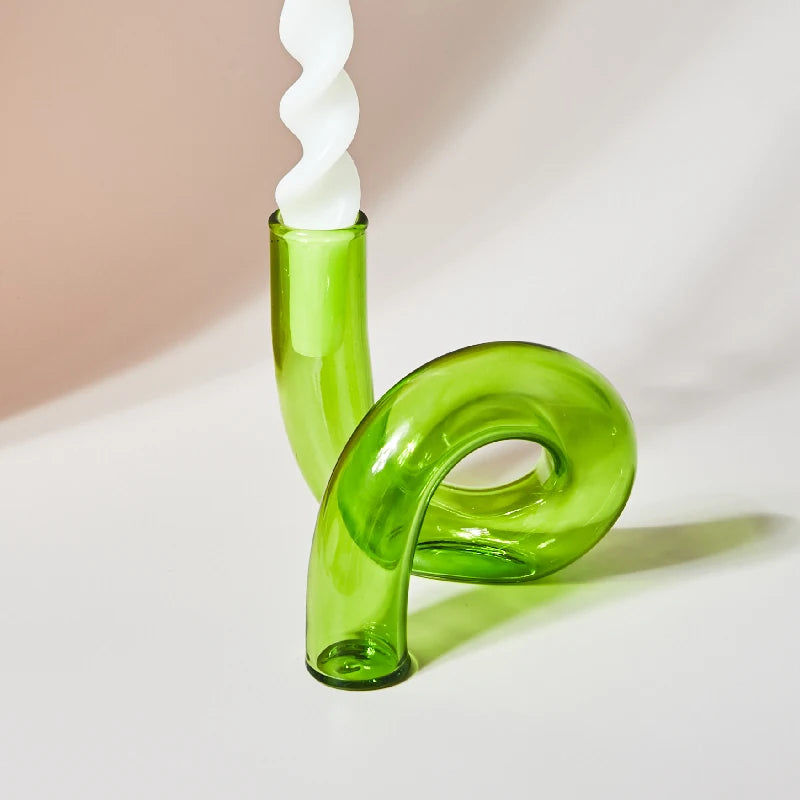 A modern, curved green glass candle holder with a twisted white candle set against a neutral background. The design features artful, fluid lines and a glossy finish.
