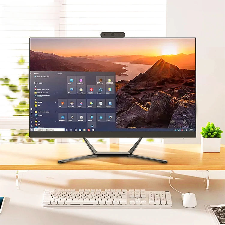 A modern computer monitor on a wooden desk with a scenic mountain sunset wallpaper. Keyboard and mouse are neatly placed underneath, with a small plant on the right side.