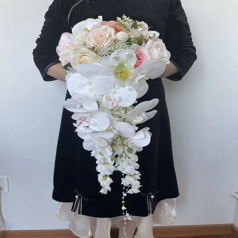 A woman in a black dress holds an elegant cascading bouquet featuring white orchids, pink and peach roses, and greenery, against a plain white background.