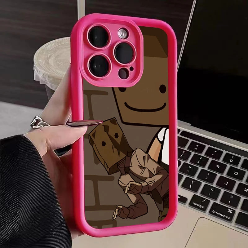 Close-up of a pink phone case with a cartoon design showing two characters wearing paper bag masks. A hand with a black sleeve holds the phone near a laptop and a cup.