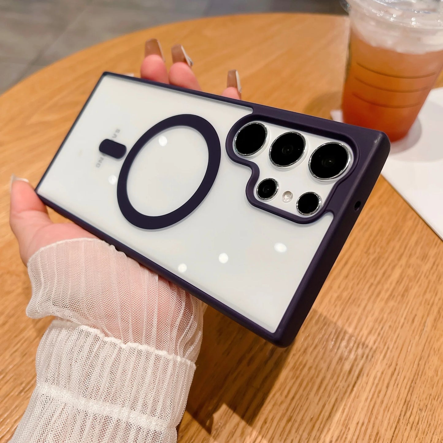 A hand with manicured nails holds a phone with a white case featuring a purple circle and multiple camera lenses. A drink sits on a wooden table in the background.