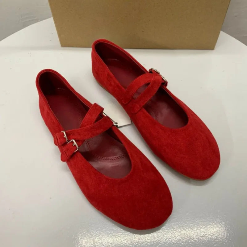 Pair of red suede Mary Jane flats with double straps and silver buckles, placed on a shiny white surface with a plain brown box in the background.