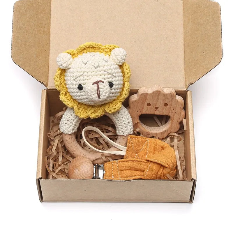 A cardboard box contains a knitted lion toy, a wooden lion-shaped teether, a pacifier clip with a wooden bead, and a yellow cloth, all nestled in shredded paper.