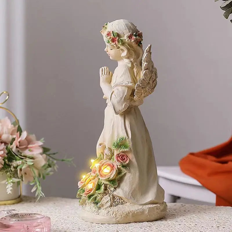 A ceramic angel figurine with floral accents and lit roses stands on a table, surrounded by soft pink flowers and a decorative orange cloth in the background.