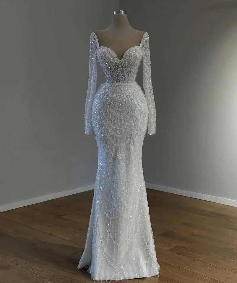 Elegant white lace gown on a mannequin with long sleeves, intricate beading, a sweetheart neckline, and a form-fitting design, displayed in a dimly lit room.