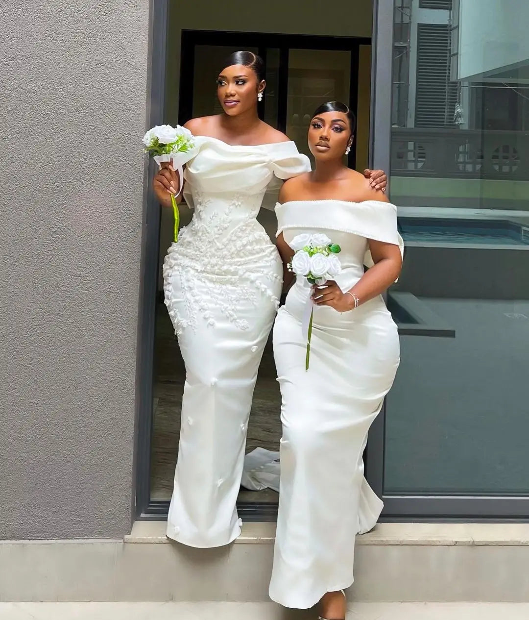 Two women in elegant white off-shoulder dresses hold small white flower bouquets. They stand close together on a modern building step, one arm around each other, exuding confidence and grace.