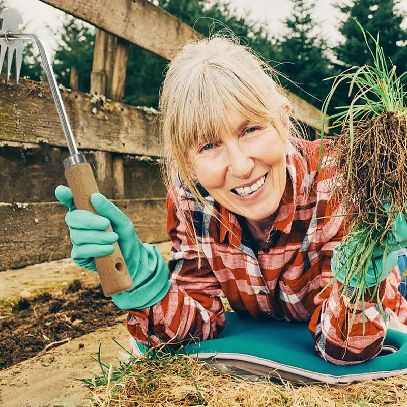 Smiling person in a plaid shirt and gloves, holding a garden tool and uprooted grass, kneels on a mat by a wooden fence. Forest background adds to the outdoor gardening scene.