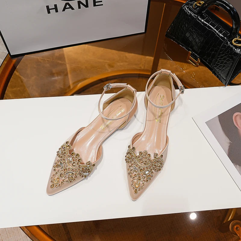 Elegant beige pointed-toe heels adorned with sparkling rhinestones and ankle straps, displayed on a table beside a black handbag and a shopping bag.