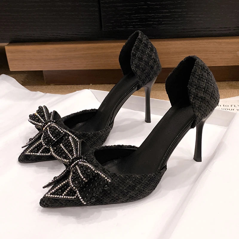 Elegant black high heels with a textured pattern, featuring pointed toes adorned with beaded bows. The shoes rest on a white surface against a dark wood background.