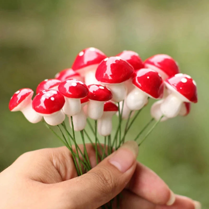 A hand holds a small bouquet of decorative red and white artificial mushrooms with green wire stems, against a blurred green background.