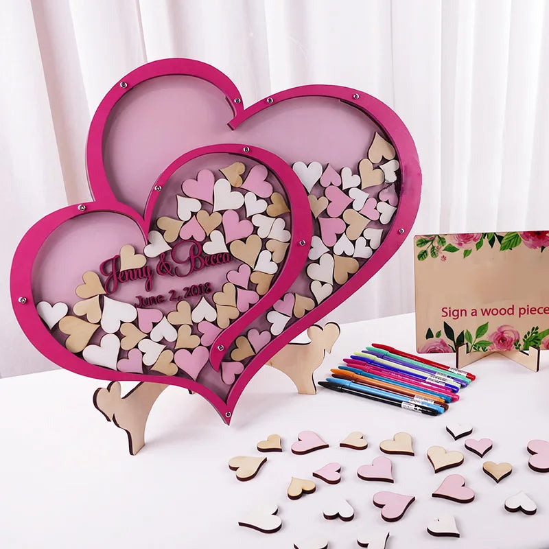 Heart-shaped wooden guestbook filled with signed pink and beige hearts. Displayed with colorful pens beside a floral sign prompting guests to write on heart pieces.