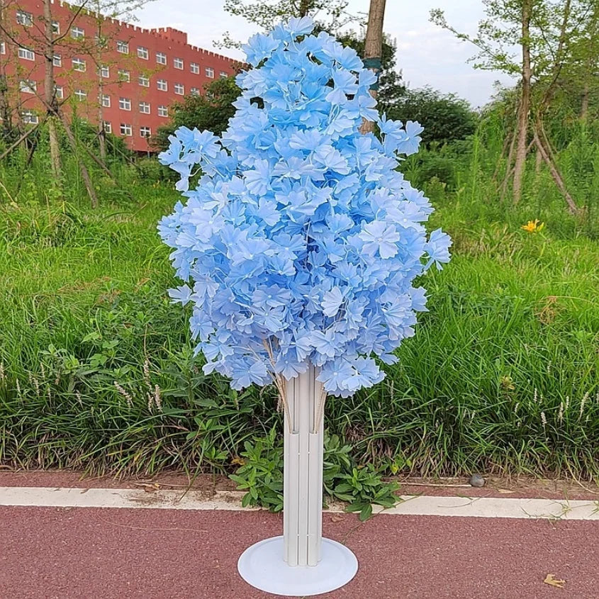 A tall decorative stand with clusters of light blue artificial flowers stands on a sidewalk, set against a backdrop of green grass and a red brick building.