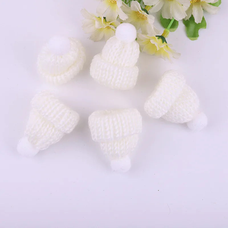 Five small, white, knitted hats with pom-poms arranged on a surface beside a bouquet of light yellow flowers.