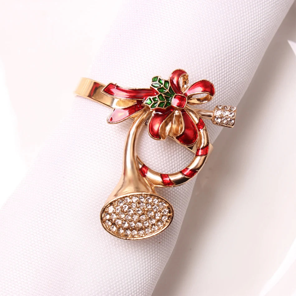 Gold napkin ring shaped like a musical horn, adorned with red and green enamel bow and holly leaves, set with small clear crystals, on a white napkin.