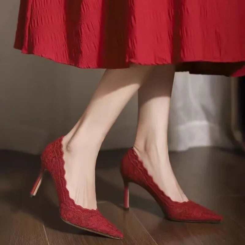 A person wearing elegant red lace high heels and a matching red skirt stands on a wooden floor, showcasing the shoes' pointed toes and scalloped edges.