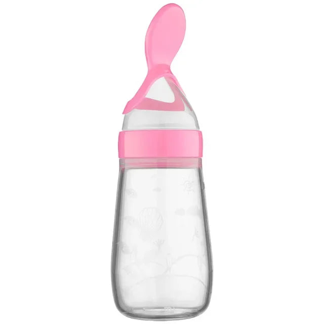 Clear baby bottle with a pink silicone spoon attachment, designed for easy feeding. The bottle is transparent, showcasing delicate embossed patterns.