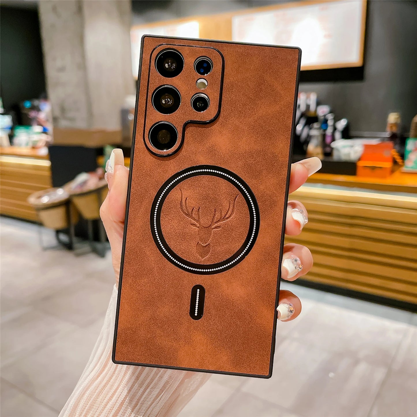 A brown leather-textured phone case featuring a deer emblem and multiple camera lenses is held up in a hand with a blurred indoor setting in the background.
