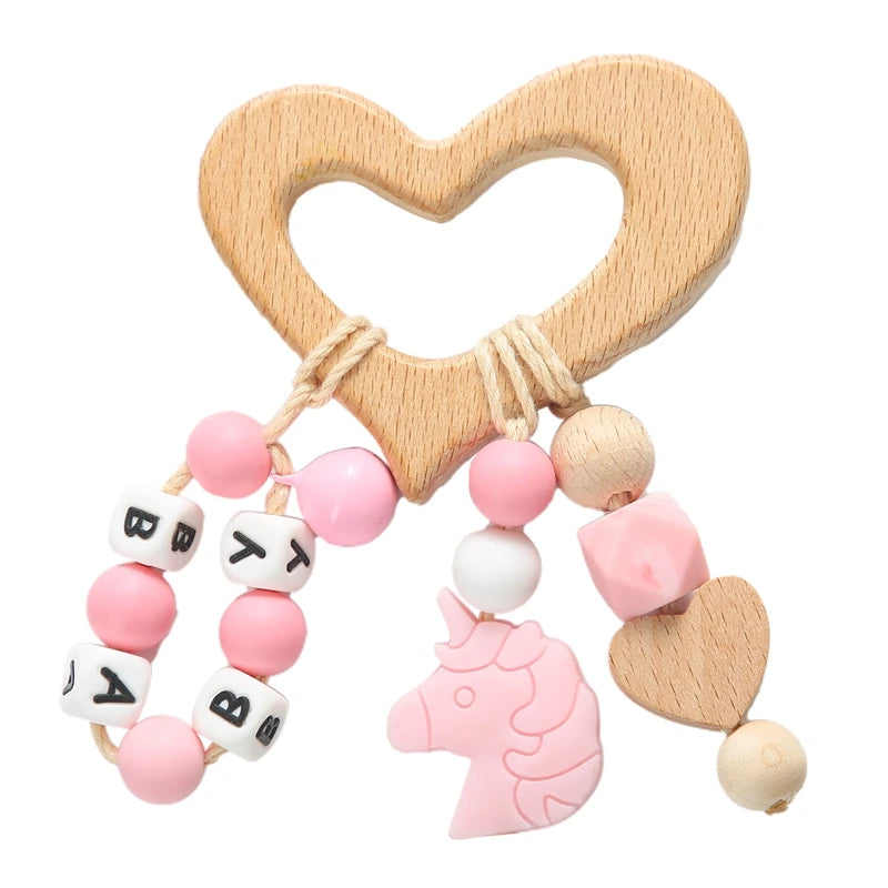 Wooden heart-shaped baby teether with pink and white beads, a pink unicorn, and a heart, featuring alphabet beads spelling "BABY" on a cord.