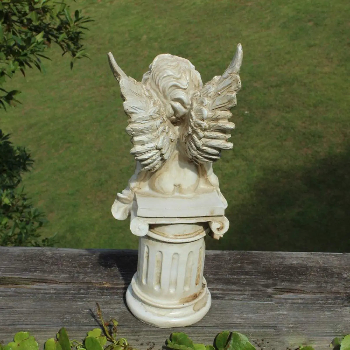 A decorative carved sculpture with intricate wings atop a detailed column, set against a grassy background and bordered by foliage.