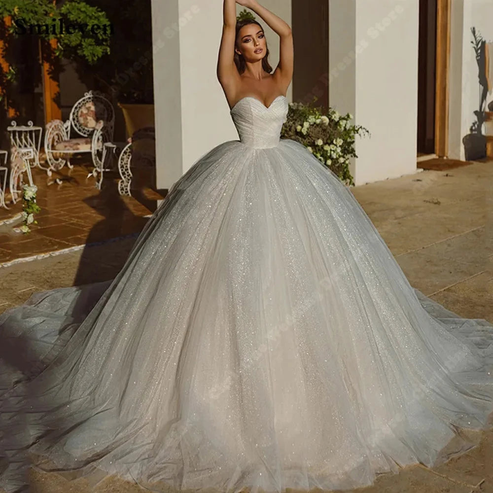 A woman poses in a sparkly A-line wedding gown with a strapless sweetheart neckline. The setting includes ornate outdoor chairs and greenery, suggesting an elegant venue.
