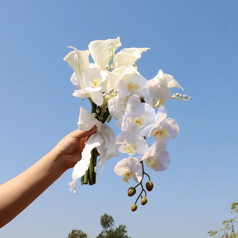 A hand holds a bouquet of white calla lilies and orchids against a clear blue sky, with a few trees visible in the distance.