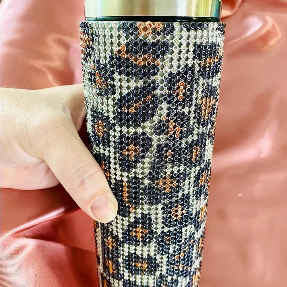 A hand holding a rhinestone-covered tumbler with a leopard print design, featuring shiny gray, orange, and black rhinestones. The background is a soft satin fabric.