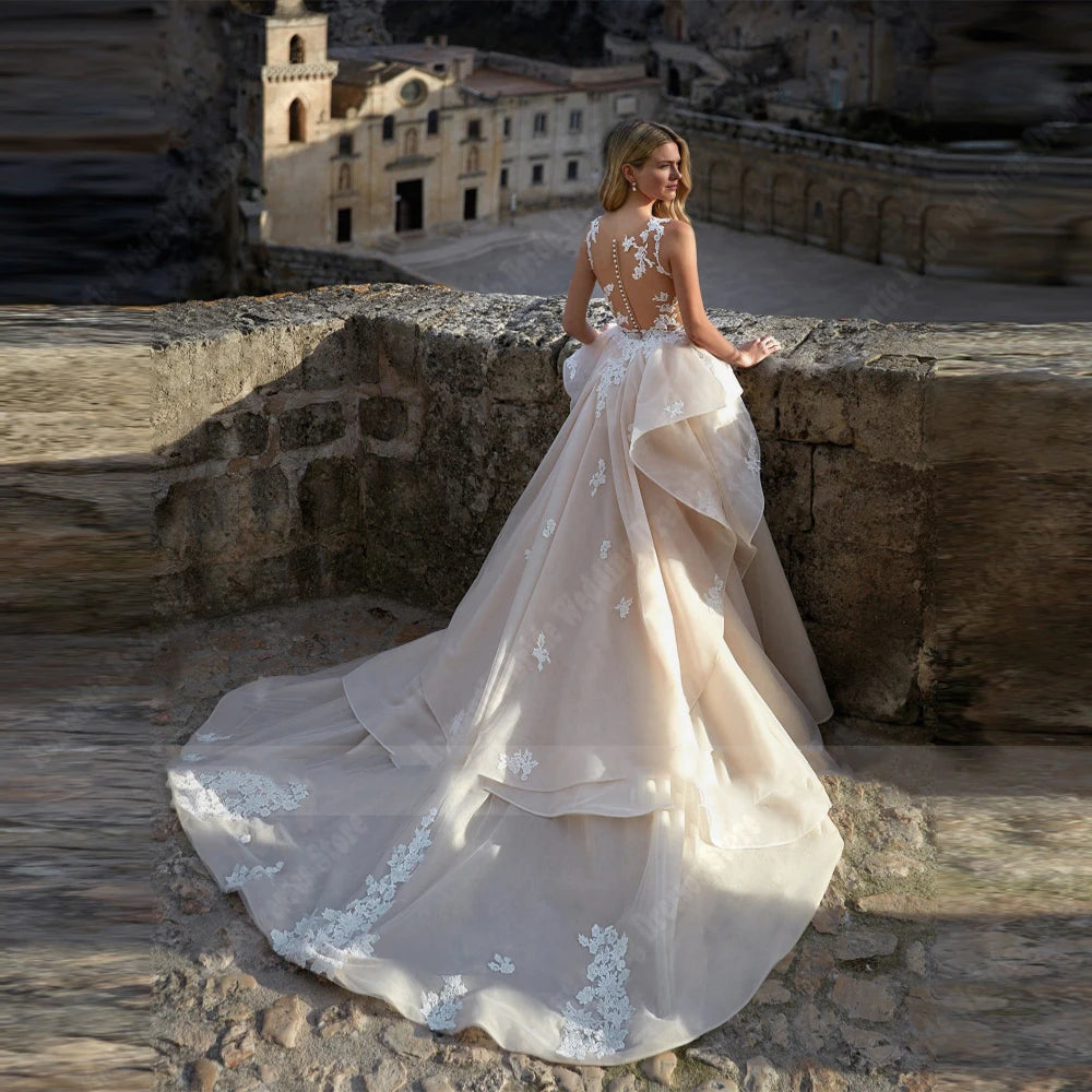 A woman in an elegant wedding dress with floral lace details and a lengthy train stands on ancient stone steps, gazing toward a historic building in the background.