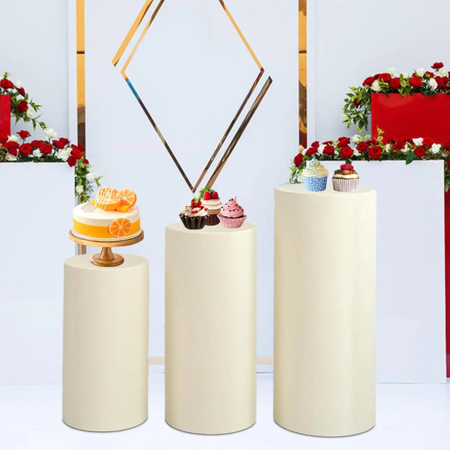 Three cream-colored cylindrical podiums display desserts: an orange-topped cake on the left, assorted cupcakes in the center, and more cupcakes on the right. Red floral decor in the background.