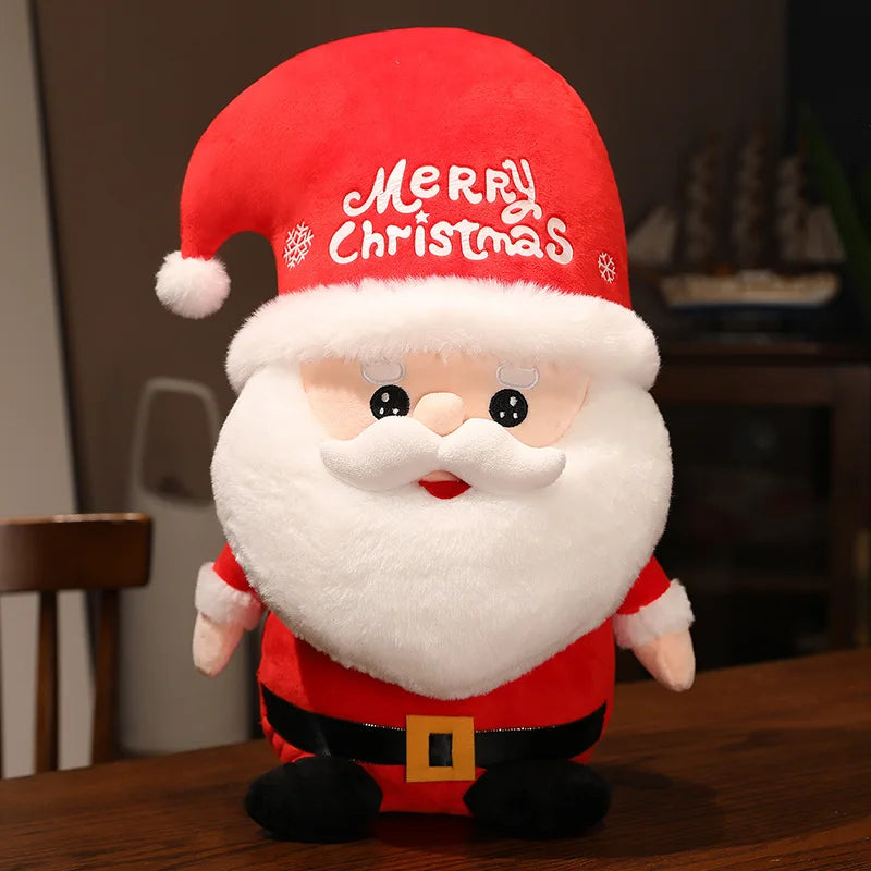 Plush Santa Claus toy with a fluffy white beard, red suit, black belt, and hat with "Merry Christmas" text. The toy is seated on a wooden table, indoors.