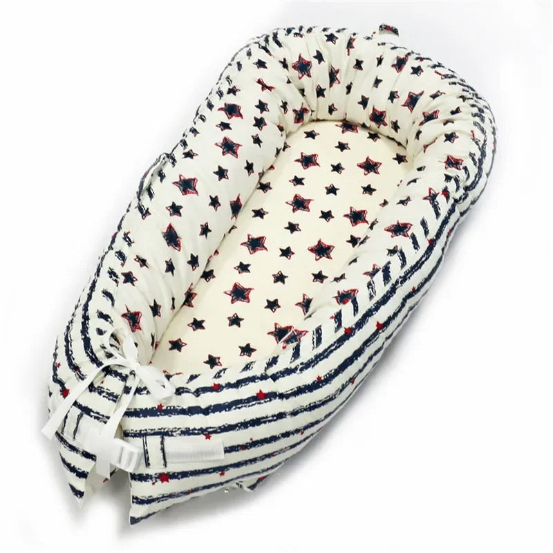White baby nest with navy blue and red star and stripe patterns. The nest has padded sides and a tied bow at the bottom, providing a cozy, secure space for a baby.