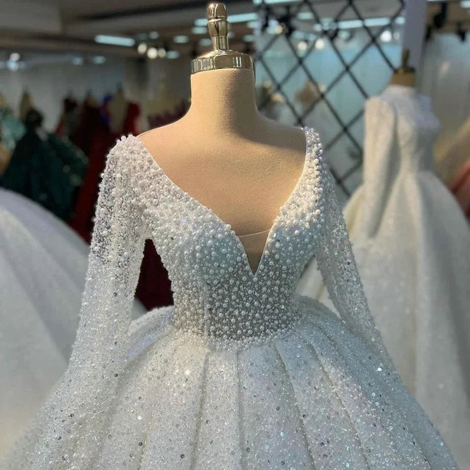 Elegant wedding dress on a mannequin, featuring a deep V-neckline with intricate beading and pearls. The dress is displayed in a boutique with other dresses in the background.