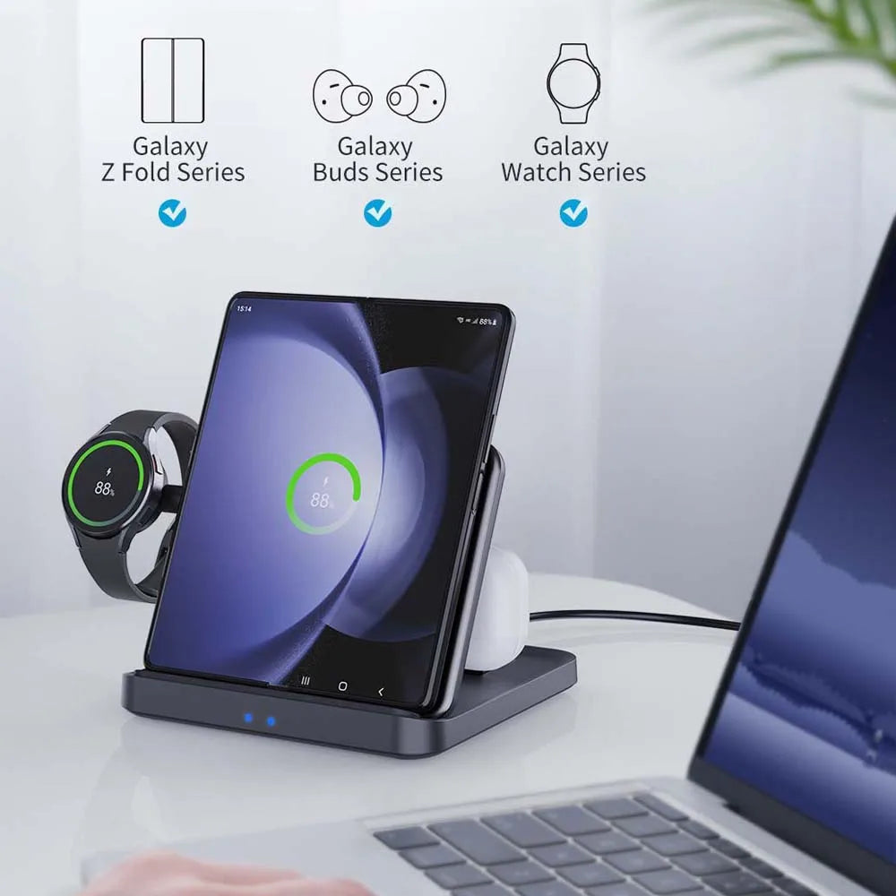 A multi-device charging stand holds a Galaxy Z Fold phone, Galaxy Watch, and Galaxy Buds, all showing charging status. Nearby is a laptop on a white desk.
