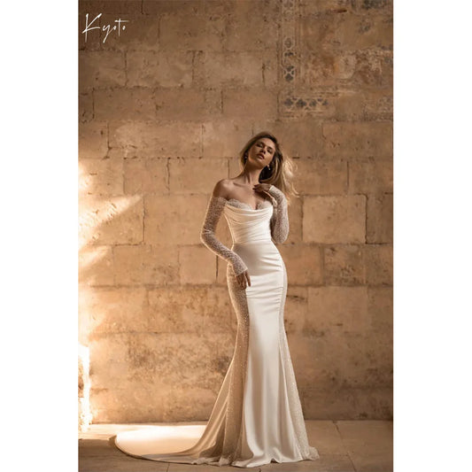A woman in a form-fitting off-shoulder lace wedding gown stands in front of a rustic stone wall, with sunlight illuminating the fabric’s shimmer and a subtle train on the floor.